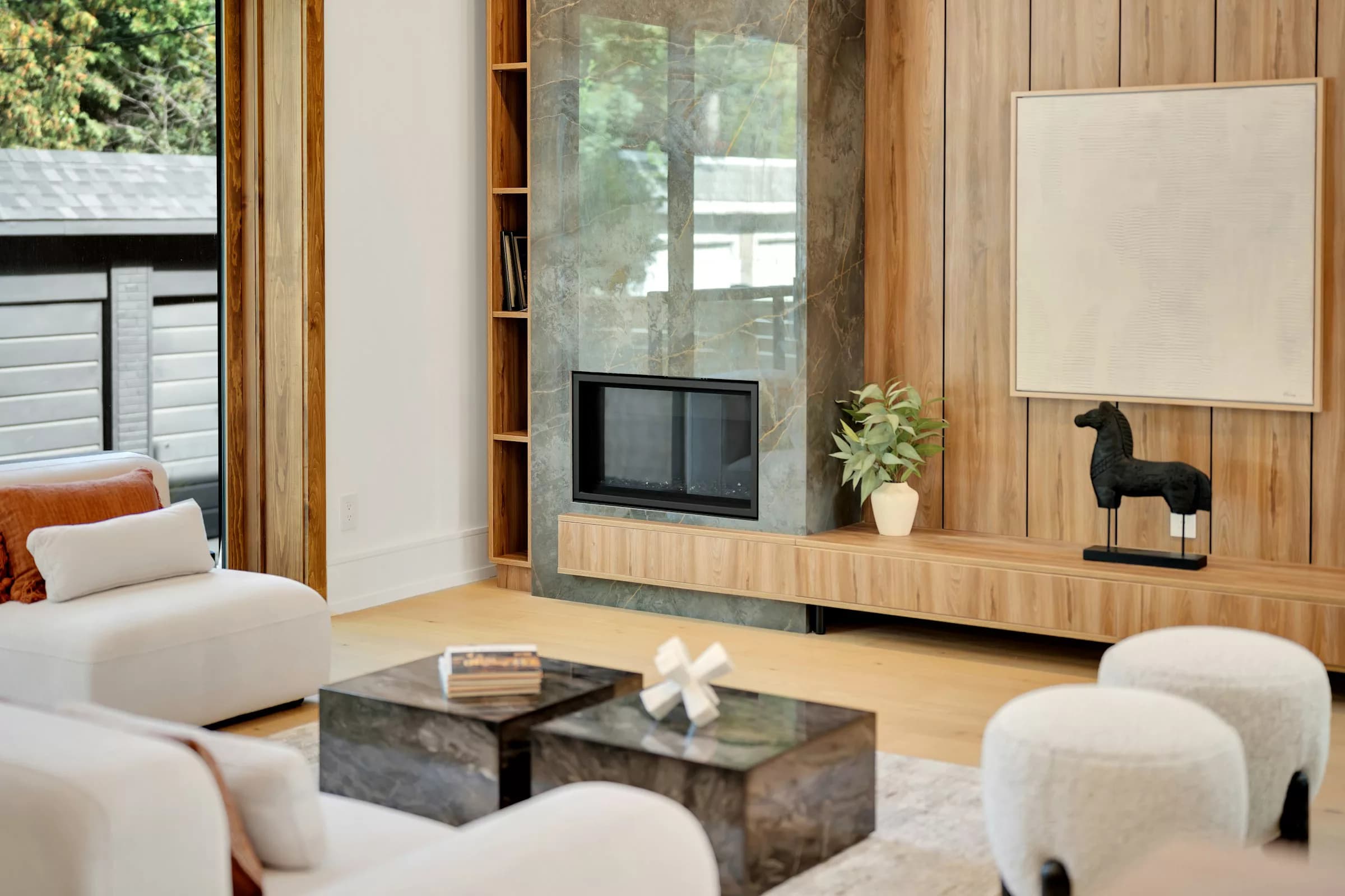 A bright, modern living room with a marble fireplace, wood paneling, and natural light from a large window